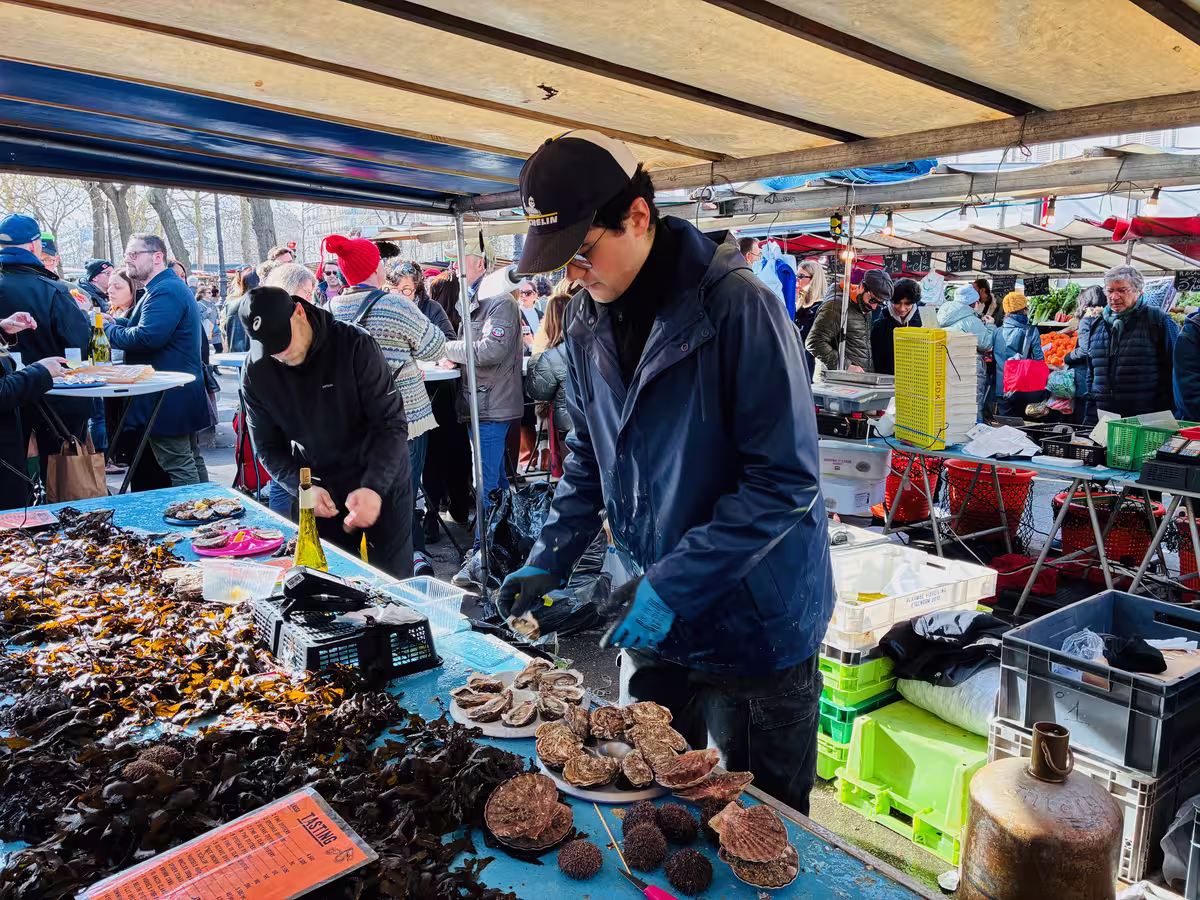 巴黎巴士地市场 (Marché Bastille) 忙碌的生蚝摊主正在开壳,背景中游客正尽情享用新鲜海鲜。